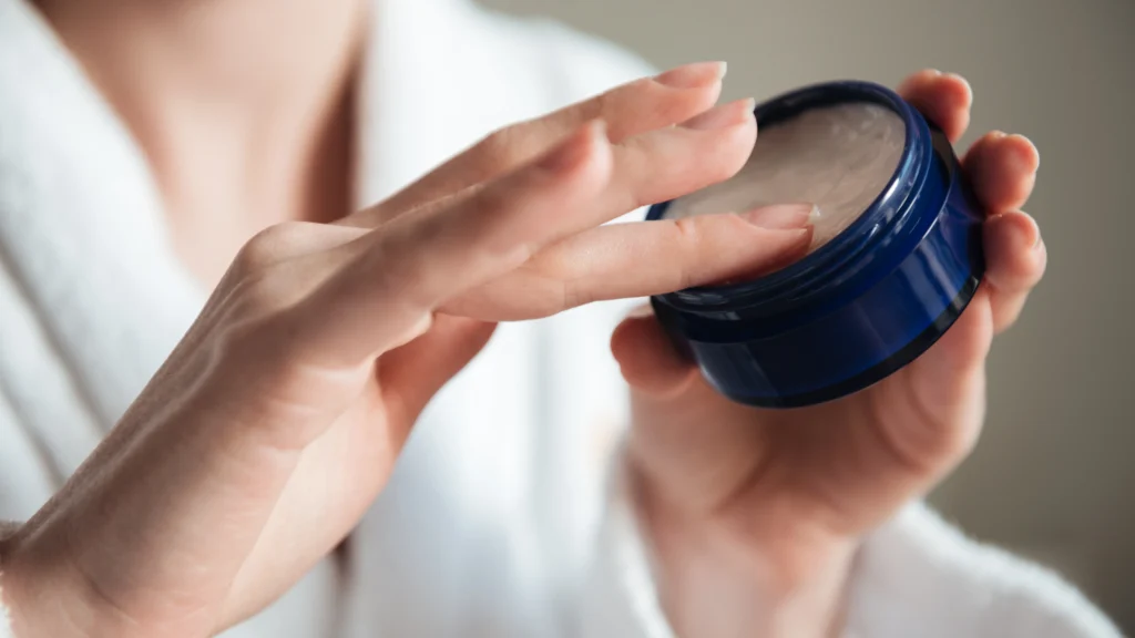 A girl holding a blue jar of skincare cream, using their finger to scoop out some product, demonstrating the application process to rehydrate skin after she follows the steps How to Get Nail Glue Off Skin Painlessly