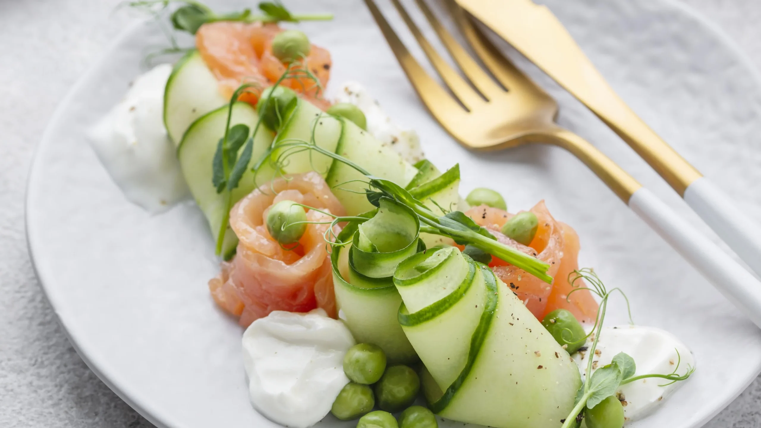 A beautifully presented low-carb salad featuring thinly sliced cucumbers wrapped around smoked salmon, garnished with fresh pea shoots and a dollop of creamy yogurt, accompanied by a gold fork on a minimalist white plate