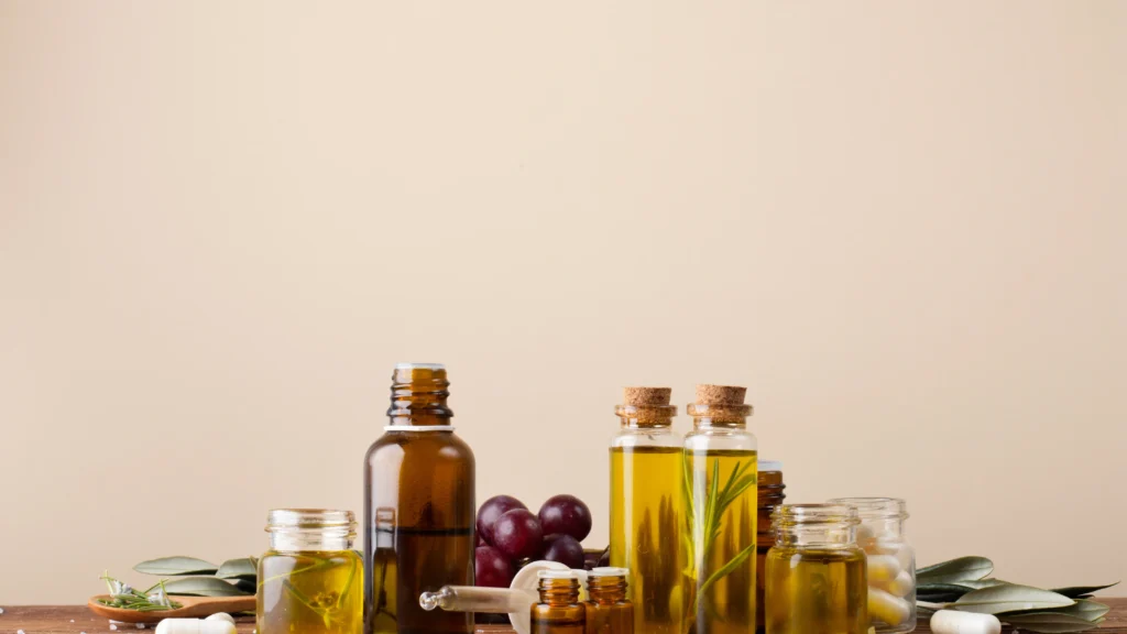 A collection of essential oils and natural ingredients, including bottles of various sizes filled with oils, fresh grapes, olive leaves, and scattered salt crystals, arranged on a wooden surface against a neutral background