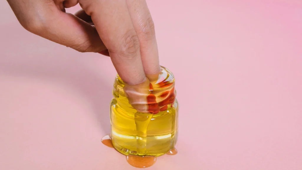 A hand dipping a finger into a small glass jar filled with golden oil, demonstrating a step in the process of how to remove press-on nails safely, with some oil dripping onto the surface below, set against a soft pink background.