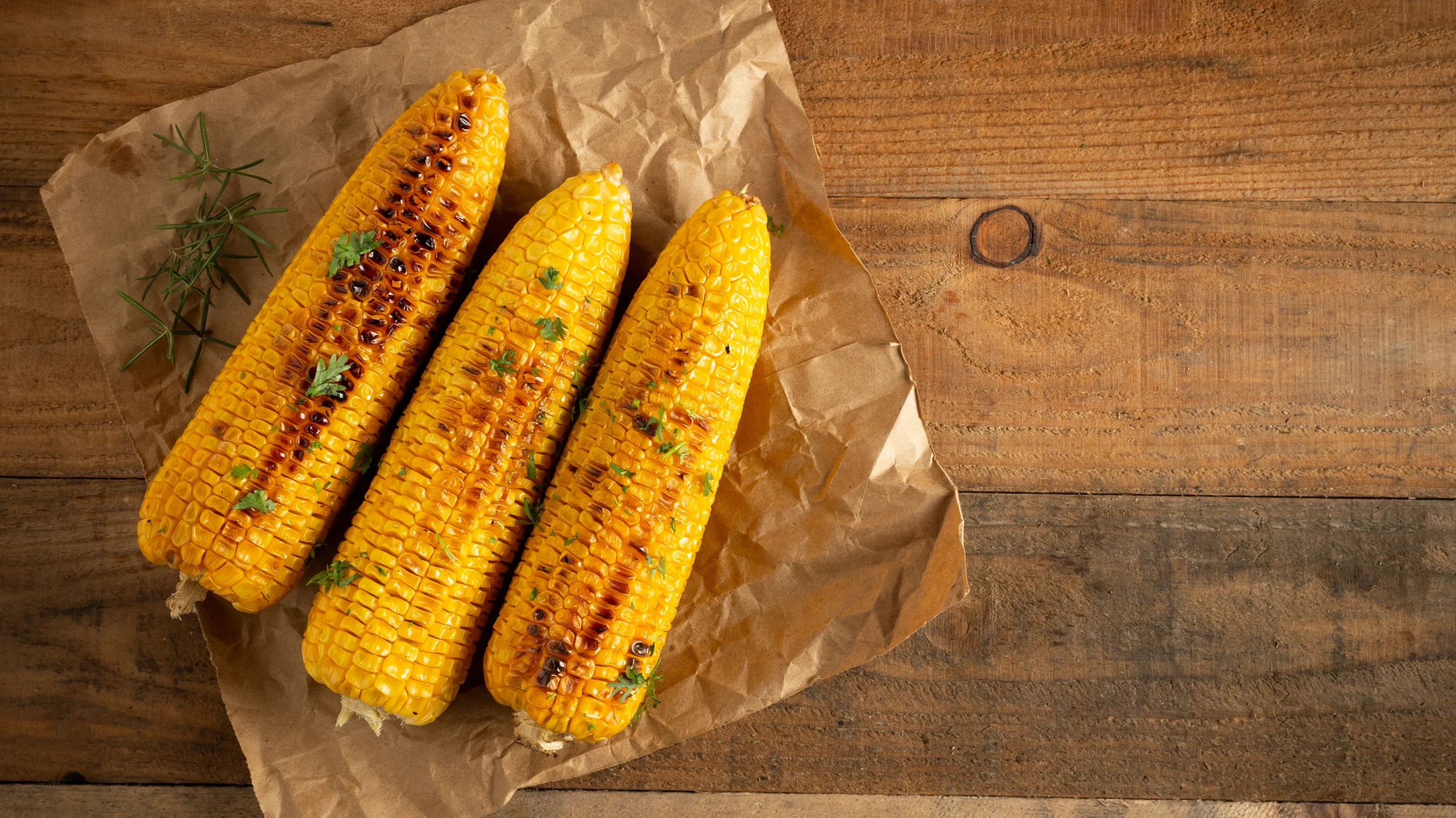 Three grilled corn cobs with charred kernels and fresh herbs, resting on brown parchment paper against a rustic wooden background, sparking curiosity about the nutritional value of corn