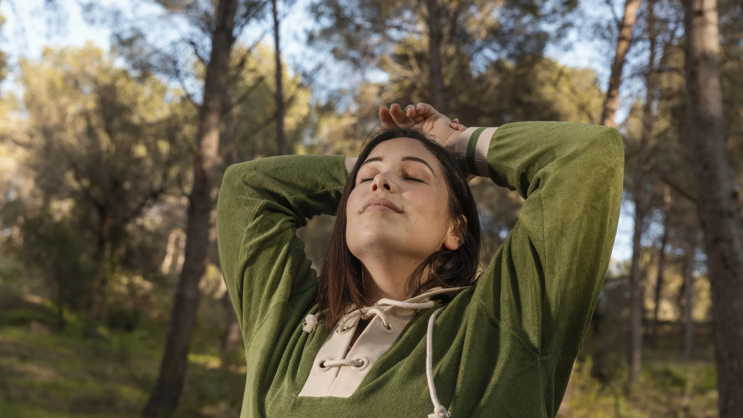 A young woman stands in a sunlit forest, her arms raised and hands clasped behind her head, eyes closed in serene contemplation—a quiet moment of connection with nature that invites reflection on 'What is mental health?' and the role of mindfulness, self-care, and tranquility in nurturing emotional well-being